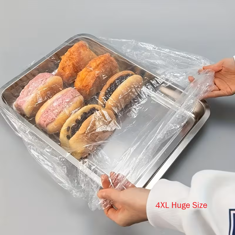 A person's hands are shown stretching a large, clear plastic food cover over a stainless steel tray containing assorted pastries and breads. The background is a clean, neutral gray studio setting.